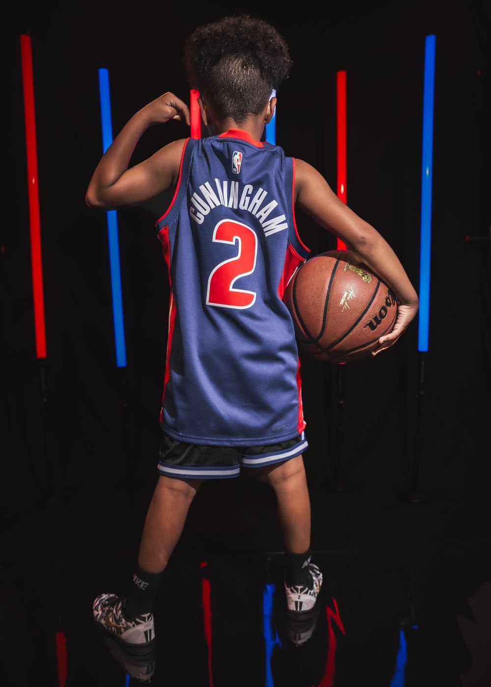Young boy wearing a Cunningham #2 Detroit Pistons NBA jersey holds a basketball in a dramatic studio setting.