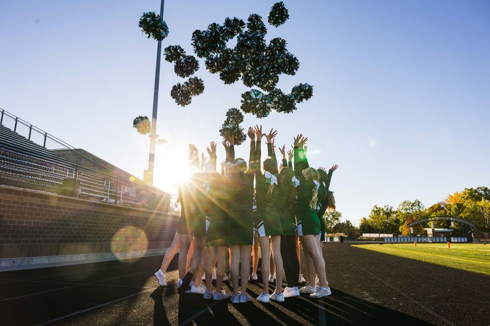 Cheerleading squad tossing green pom-poms in air during golden hour practice on stadium field.