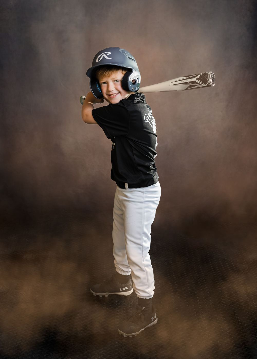 A young boy in baseball gear poses mid-swing with a bat against a dramatic studio background.