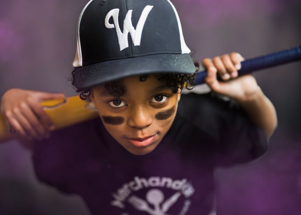 A young baseball player in a W cap holds a bat, with face paint on his cheeks in a purple smoky setting.