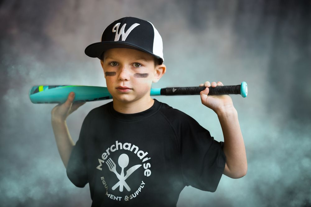 A confident young baseball player in a W cap rests a teal bat on his shoulder against a smoky background.