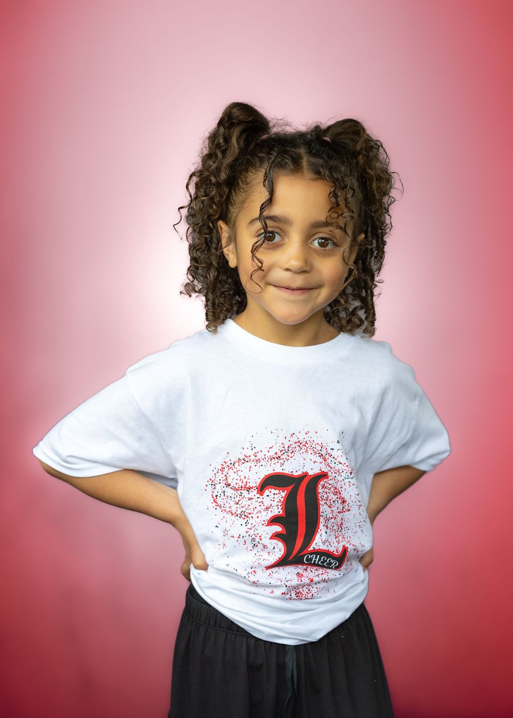 Young girl in white University of Louisville L logo t-shirt posing with hands on hips against pink background.