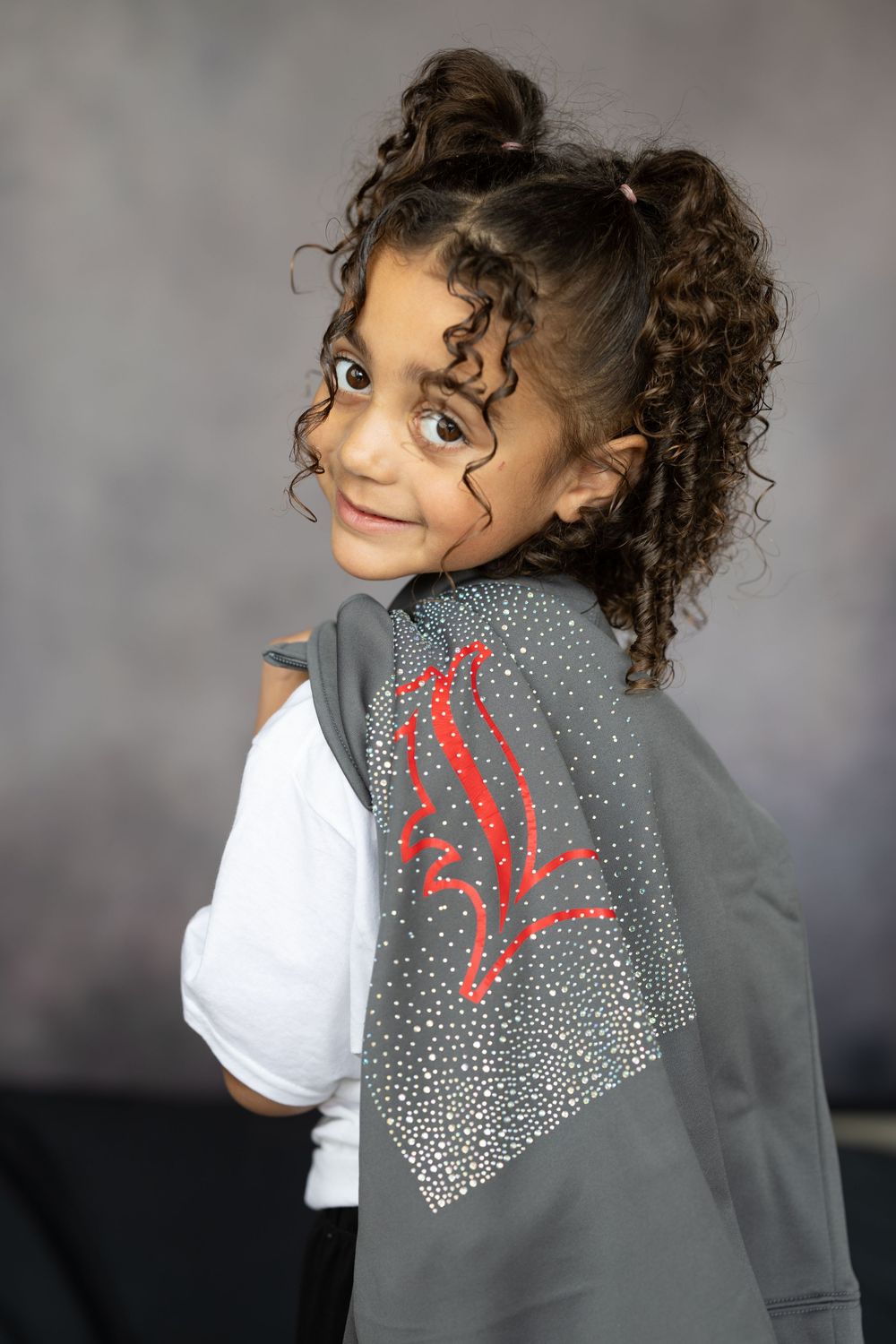 Young girl showing back of gray rhinestone L logo jacket while smiling over shoulder against gray background.