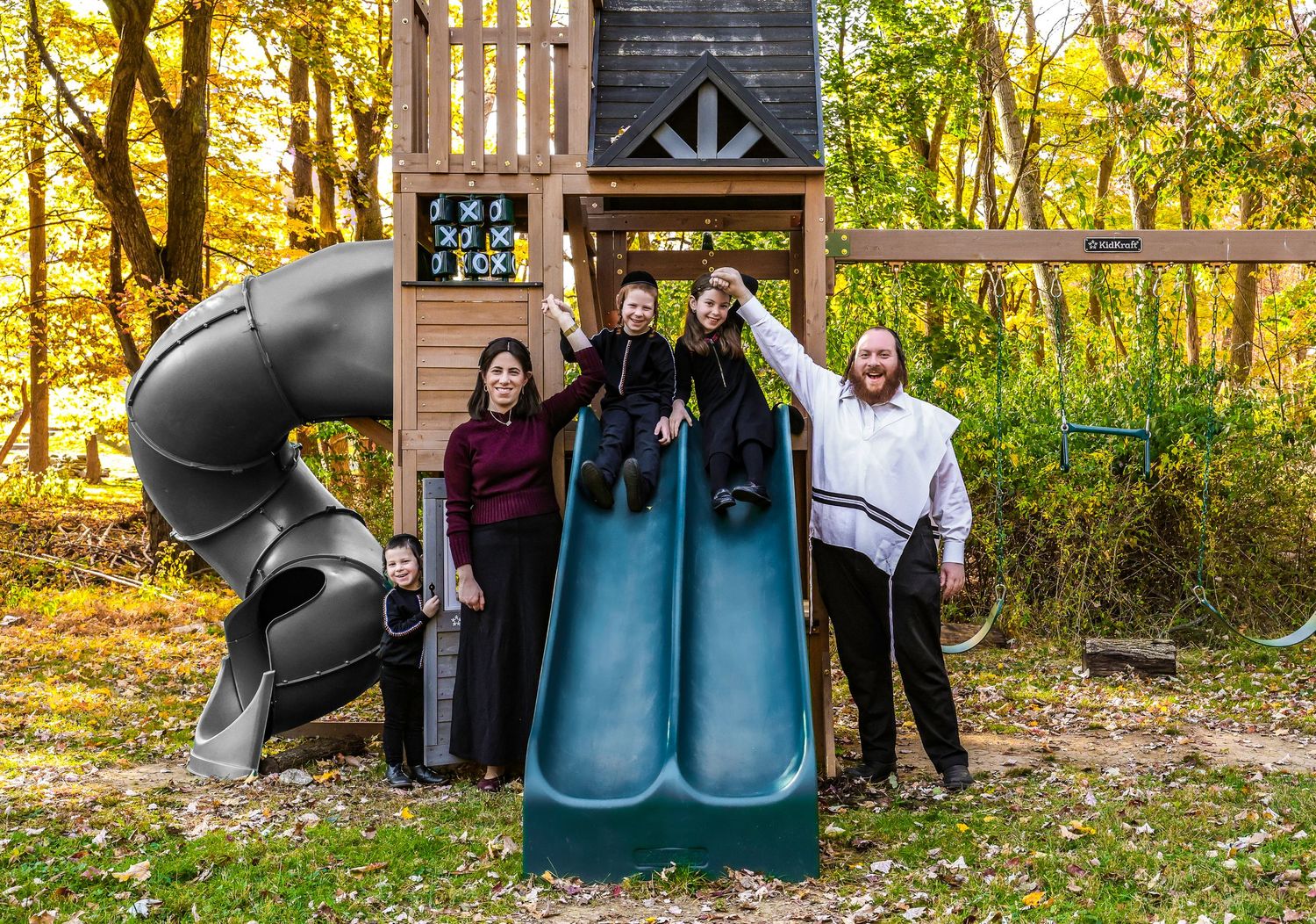 Family of five posing on a backyard playground slide surrounded by autumn foliage.