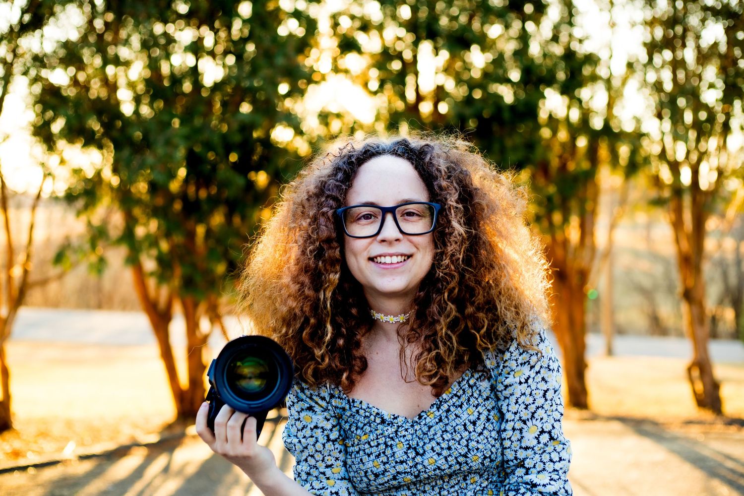 Young photographer with curly hair and glasses holds professional camera with telephoto lens in outdoor garden setting.