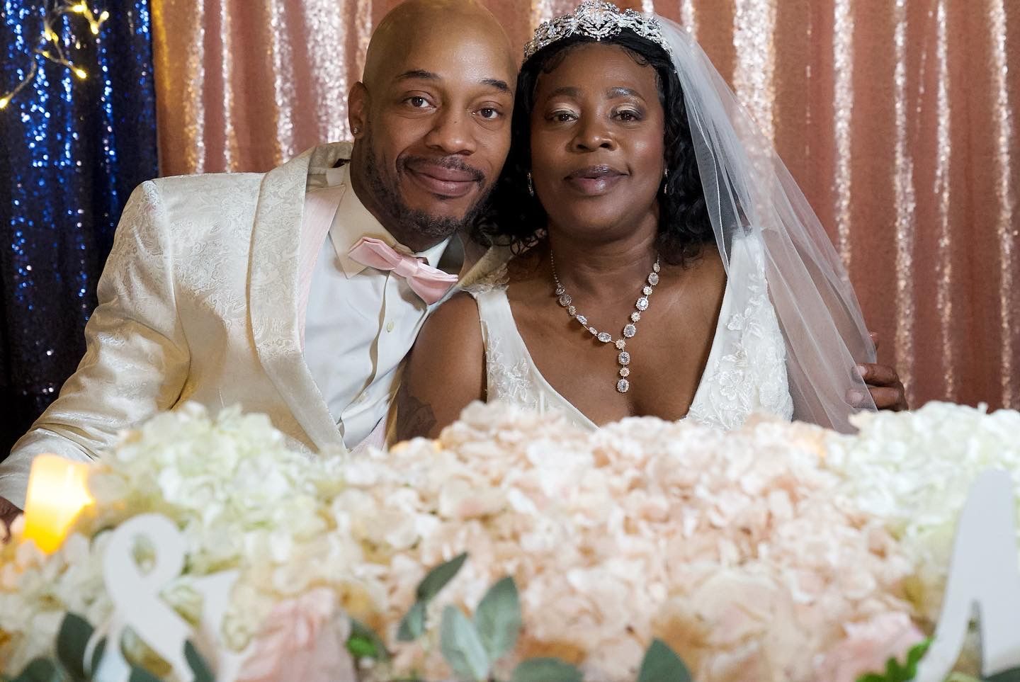 A formal wedding couple poses behind a table decorated with pink and white floral arrangements.