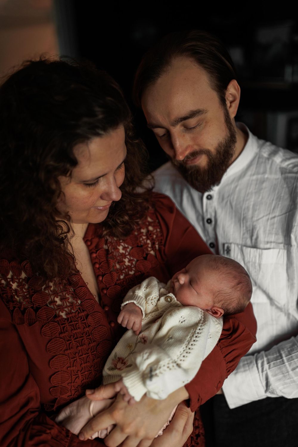 A tender family moment with a newborn baby dressed in white being held while parents gaze lovingly at their child.