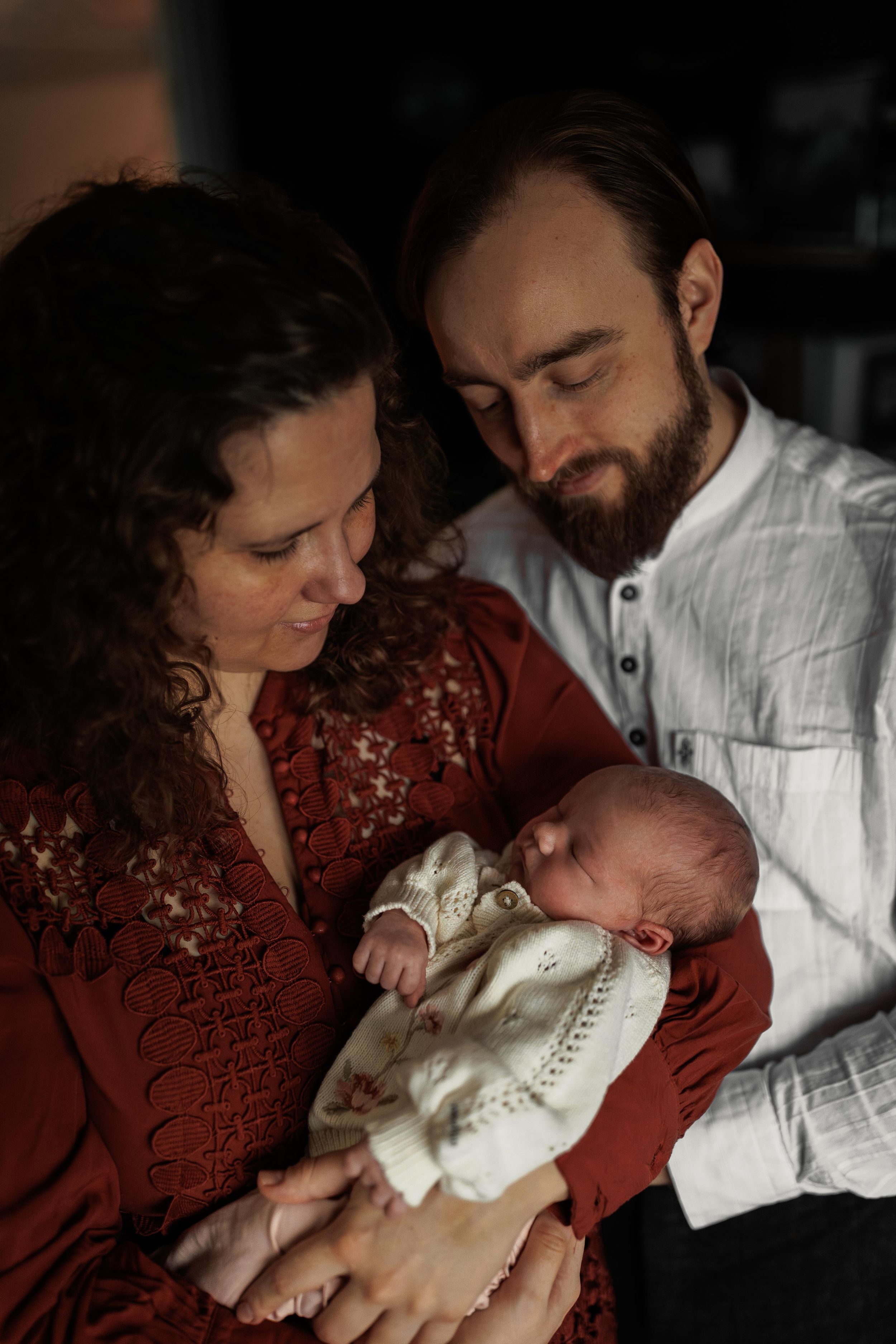 A tender family moment with a newborn baby dressed in white being held while parents gaze lovingly at their child.