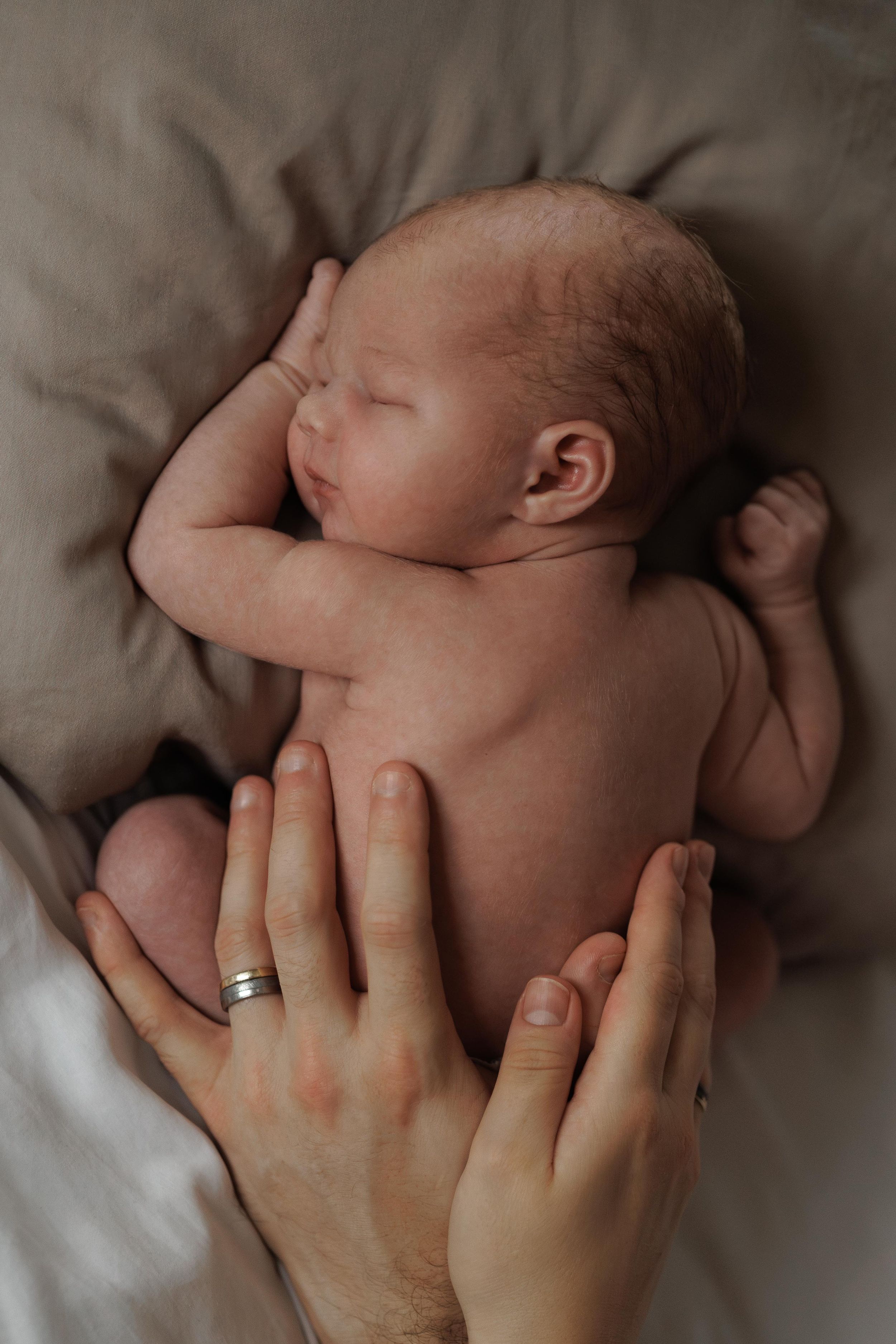 A parent's hands gently cradle their sleeping newborn baby on soft beige bedding.