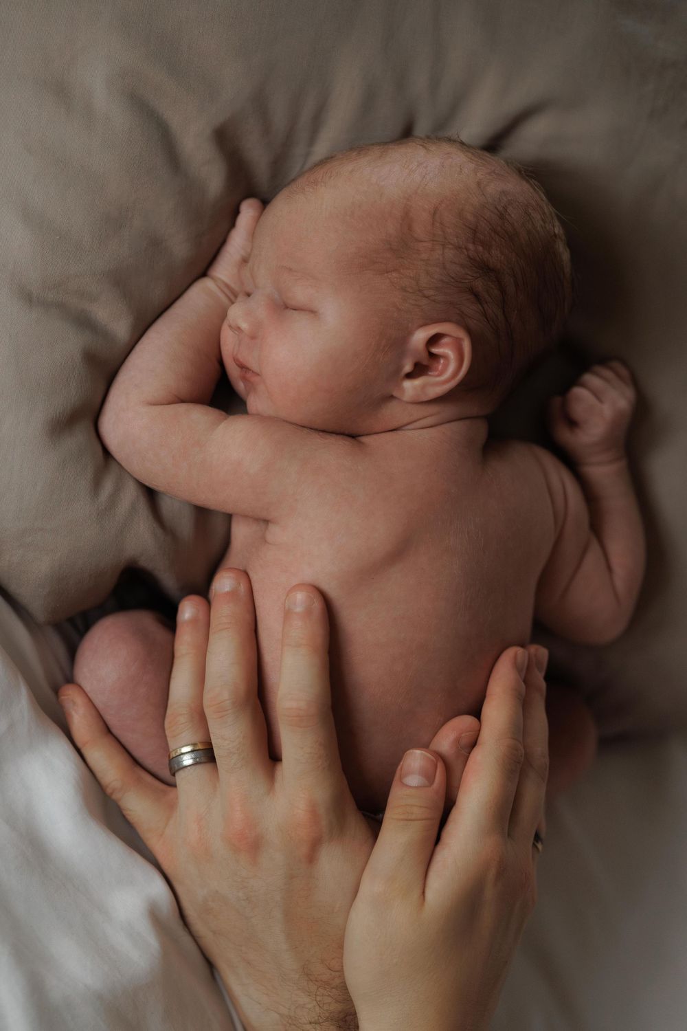 A parent's hands gently cradle their sleeping newborn baby on soft beige bedding.