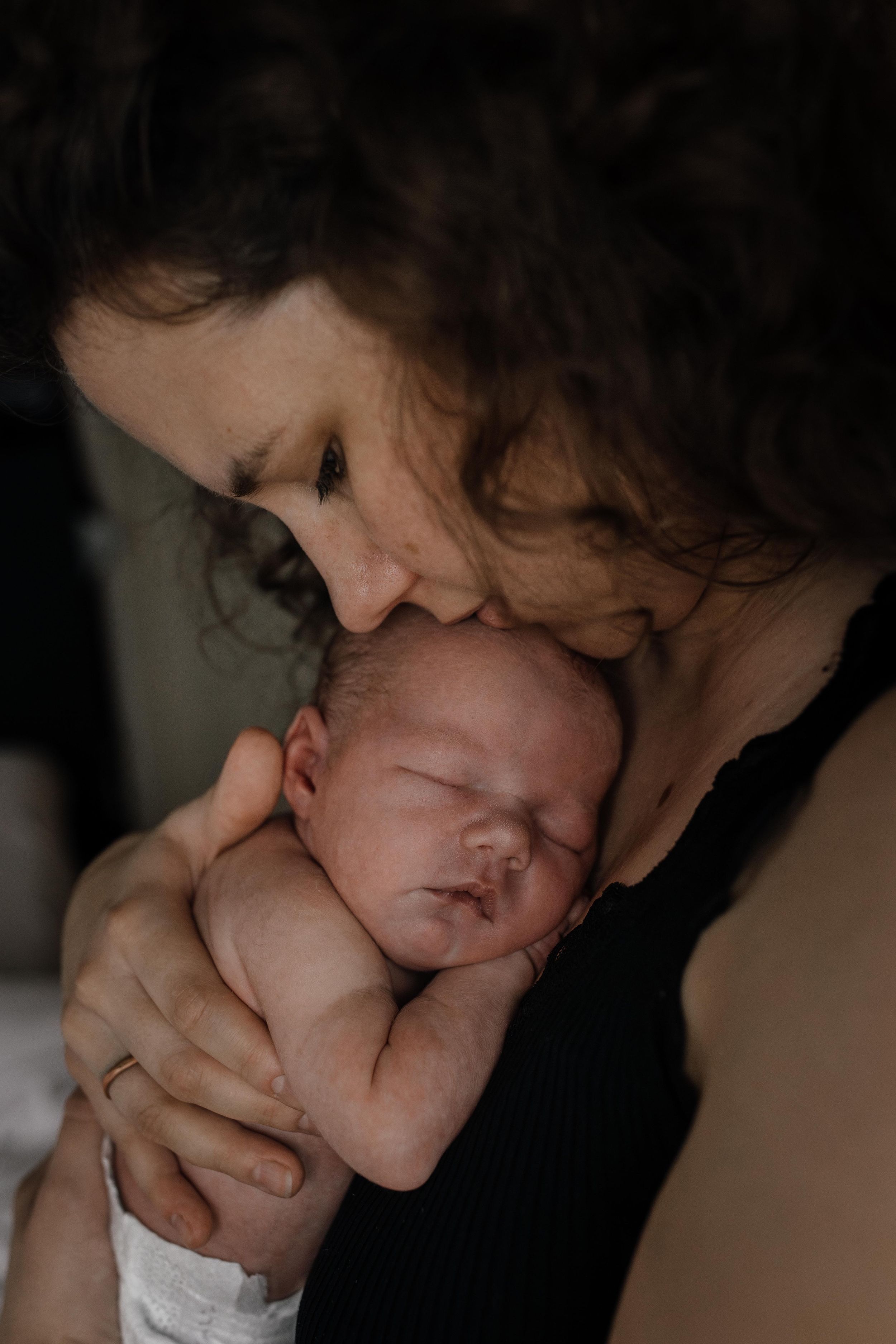 A tender moment shows a newborn baby being cradled against someone wearing black in moody, intimate lighting.