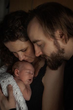 Two parents lovingly embrace their sleeping newborn baby swaddled in white lace in an intimate portrait.