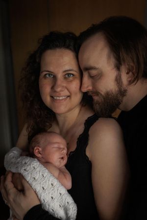 A loving couple cradles their newborn baby wrapped in a delicate white lace blanket in a dark, intimate portrait.