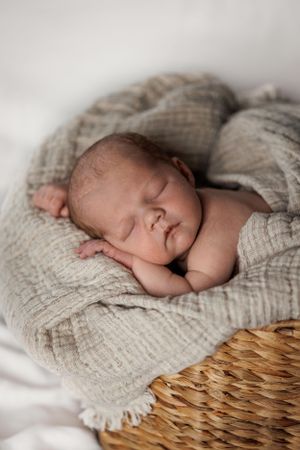 Sleeping newborn wrapped in soft beige muslin blanket resting on woven basket during professional photoshoot.