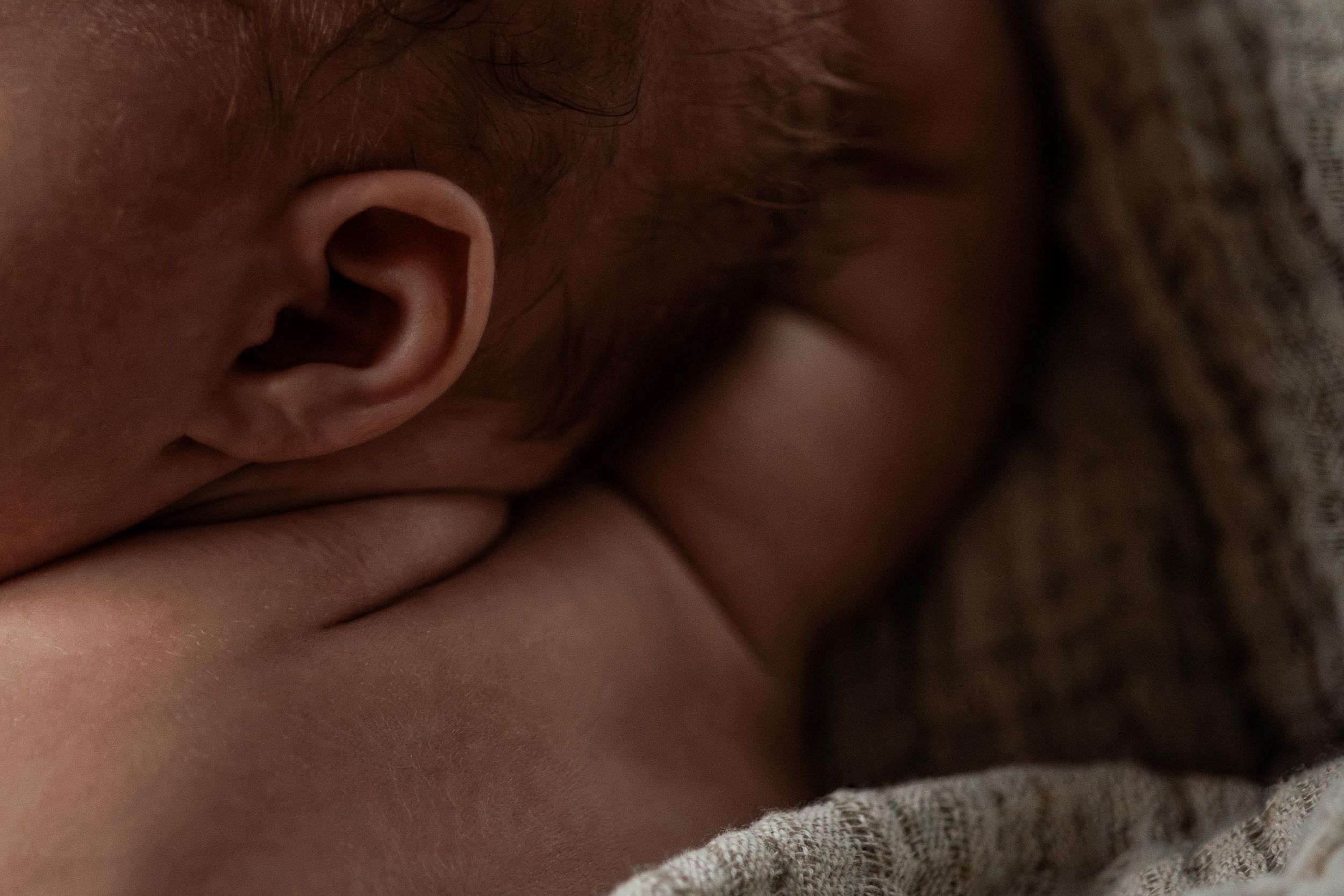 Close-up side view of a baby's ear and cheek against a soft gray textile background.