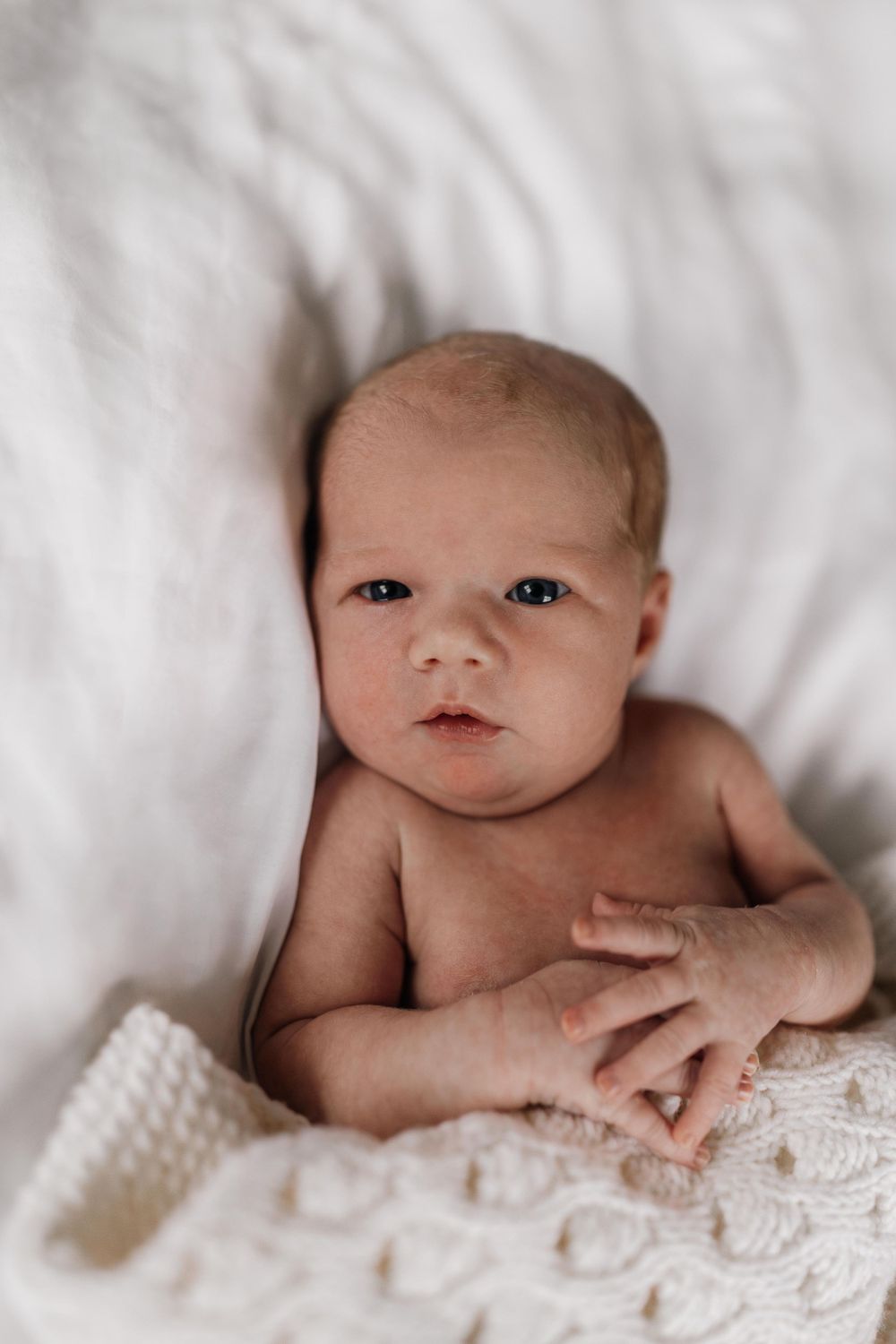 Sweet newborn baby lying on white textured blanket with soft lighting in intimate portrait setting.