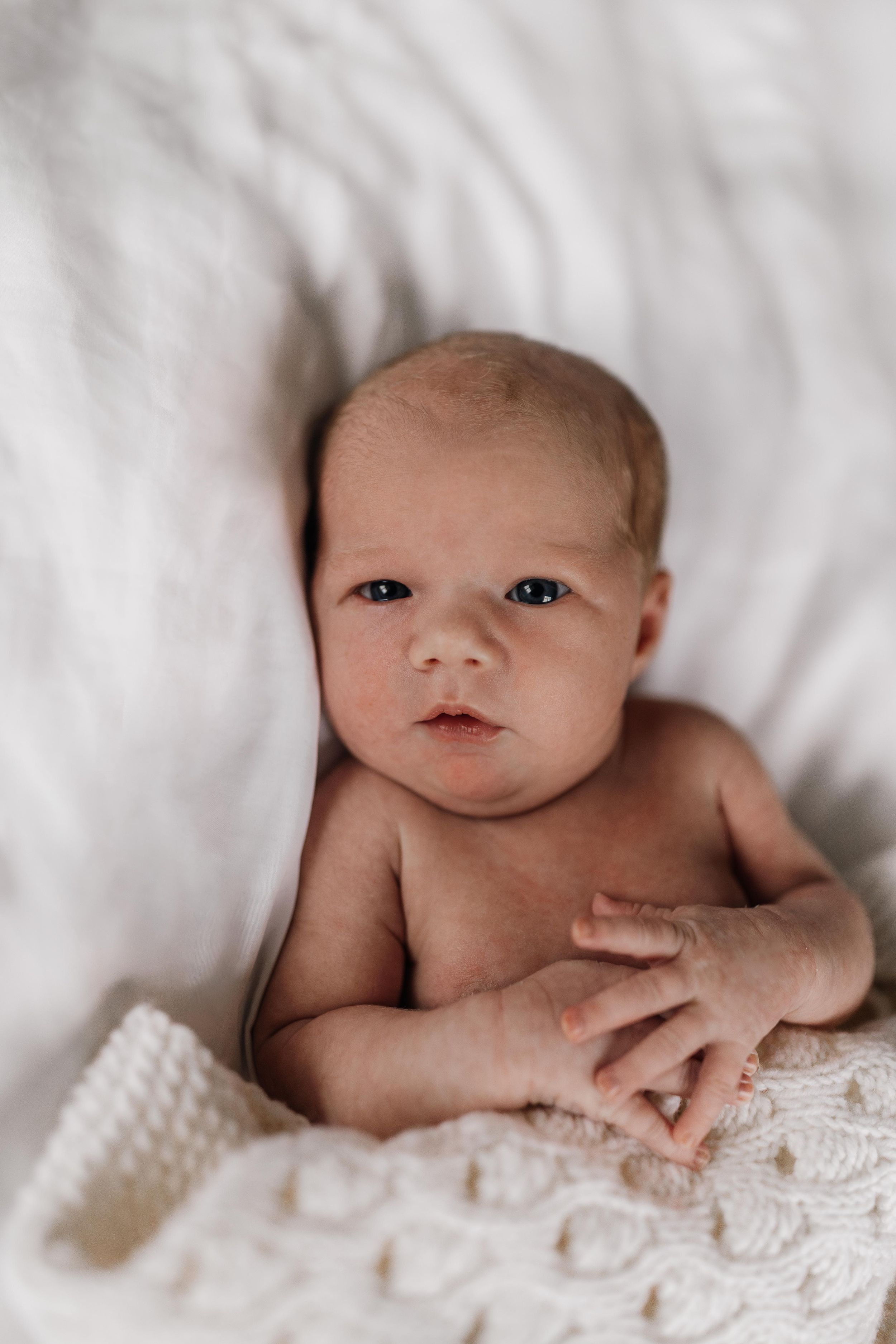 Sweet newborn baby lying on white textured blanket with soft lighting in intimate portrait setting.