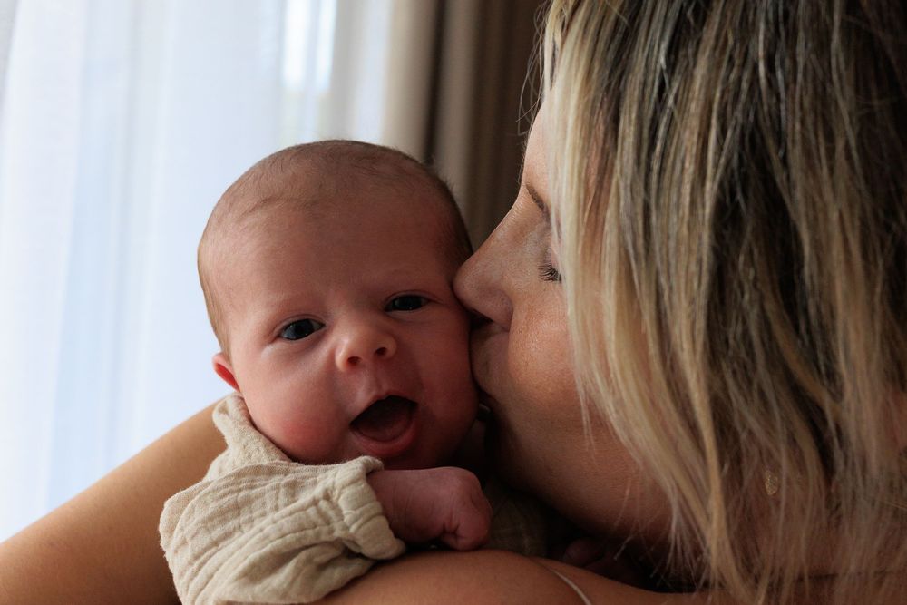 A tender moment between mother and newborn baby as she gives a loving kiss while holding them by a window.