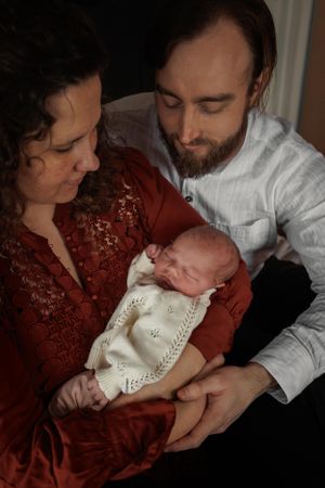 A couple in red and white clothing tenderly hold their sleeping newborn baby wrapped in a knitted white blanket.