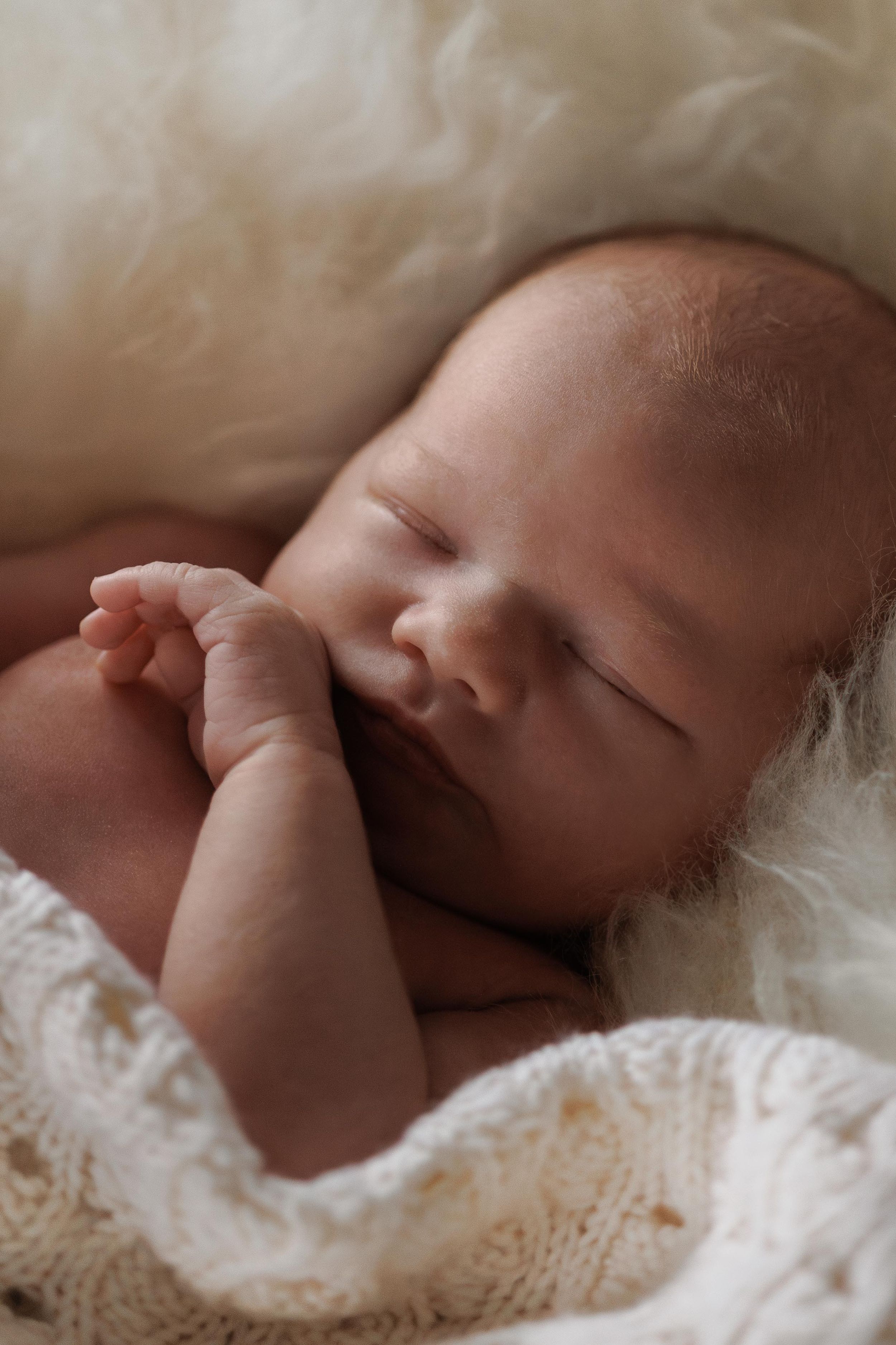 A peaceful sleeping baby wrapped in soft white knitted blankets rests on cream-colored bedding.