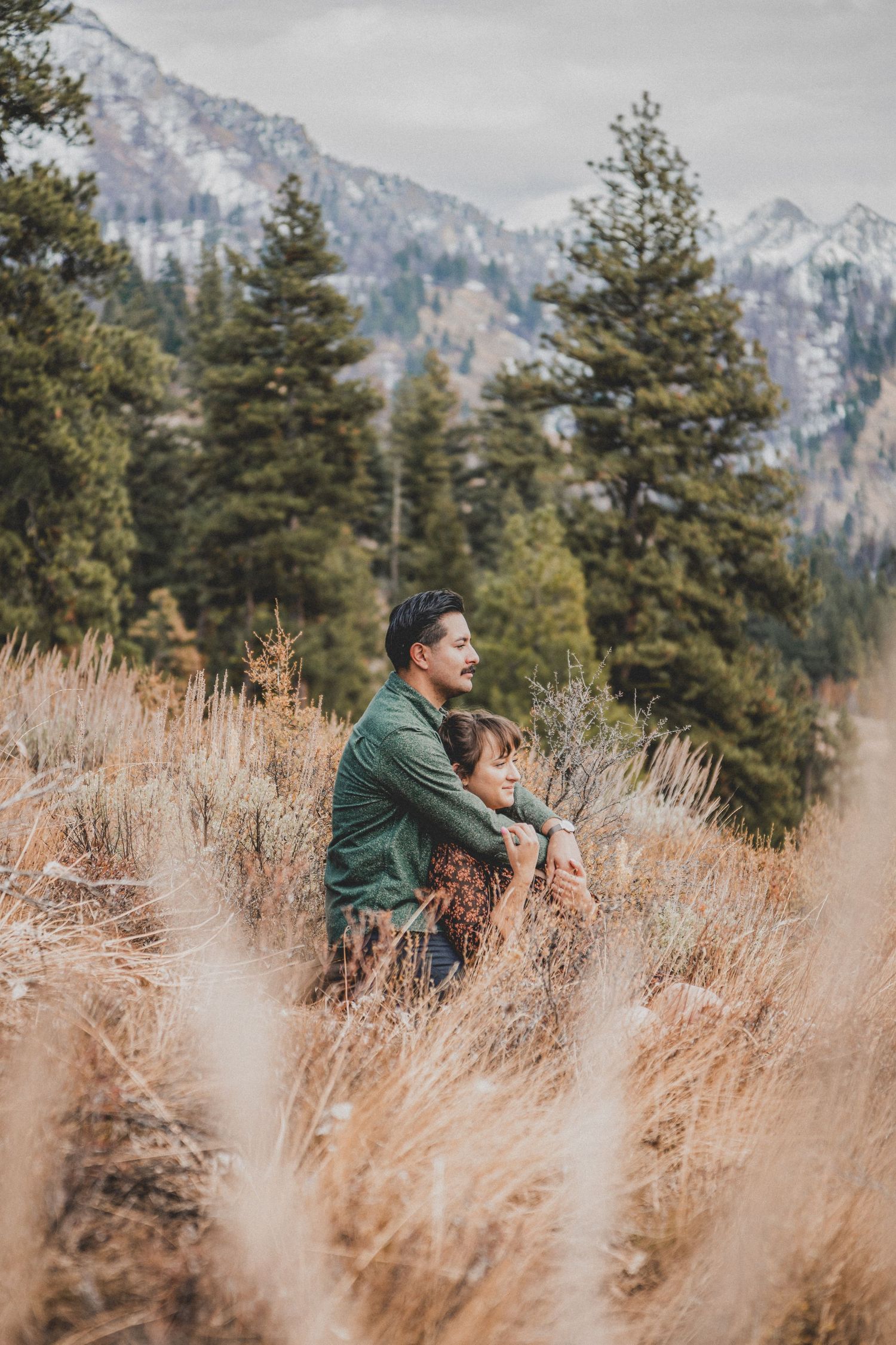 Two people stand together in tall dried grass with evergreen trees and snow-capped mountains in the background.