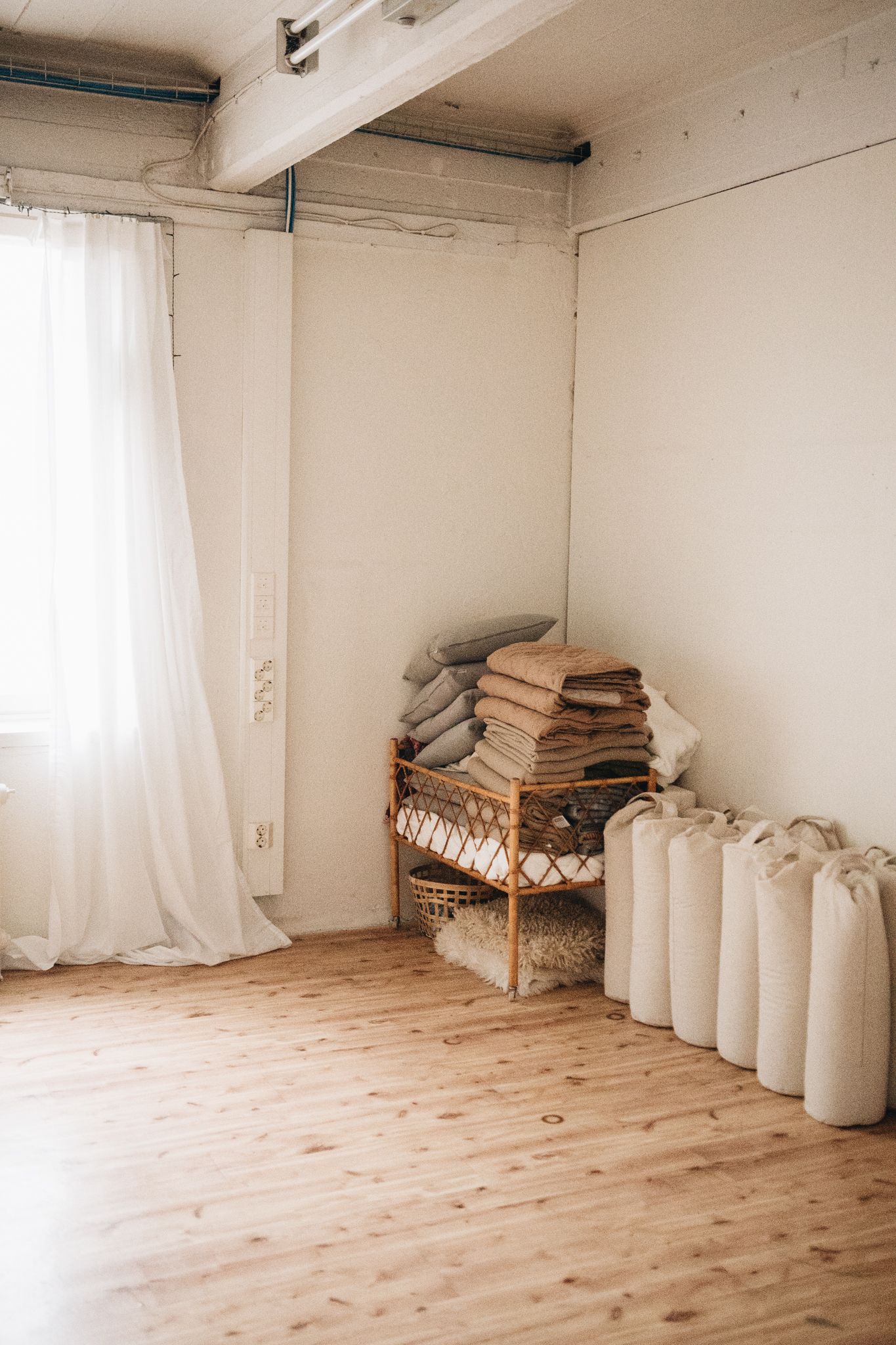 Minimalist interior with white curtains, wooden floor, and stacked linens on a bamboo bench against white walls.