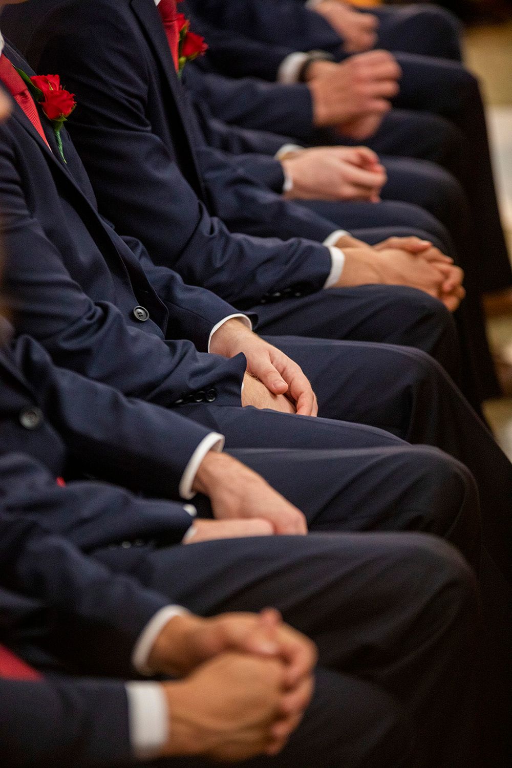 Row of groomsmen in navy suits with red boutonnieres sitting formally.