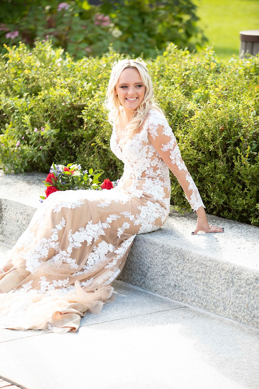 Bride in lace dress poses gracefully on stone ledge with garden backdrop.