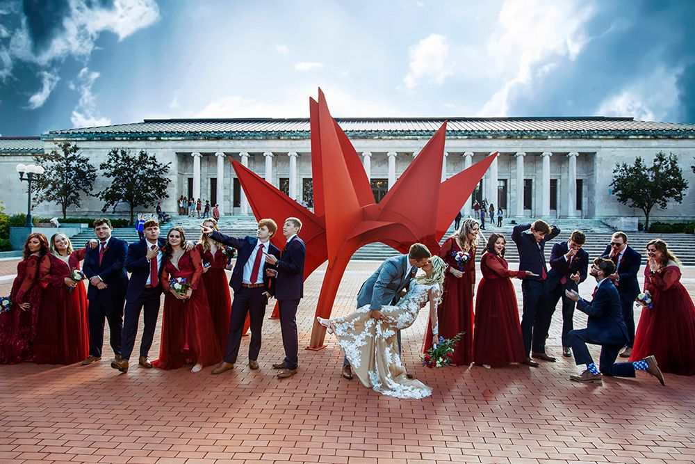 Wedding party poses with dramatic red art sculpture outside elegant building.