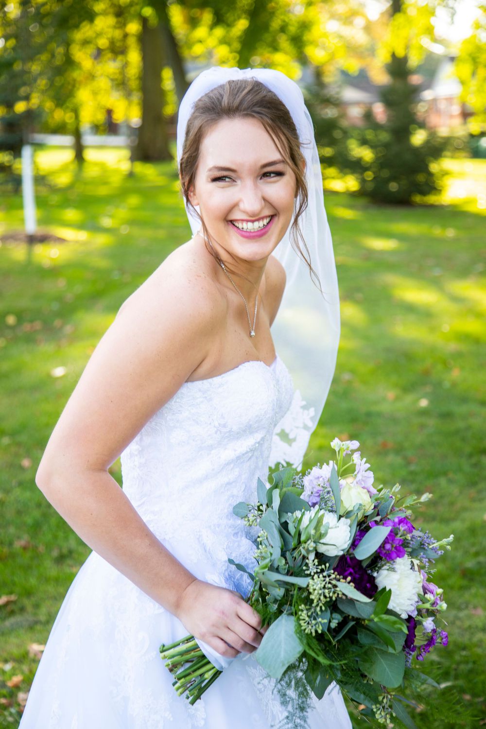 A bride wearing a white wedding dress and veil holds a purple and white bouquet while posing outdoors in a sunlit garden.