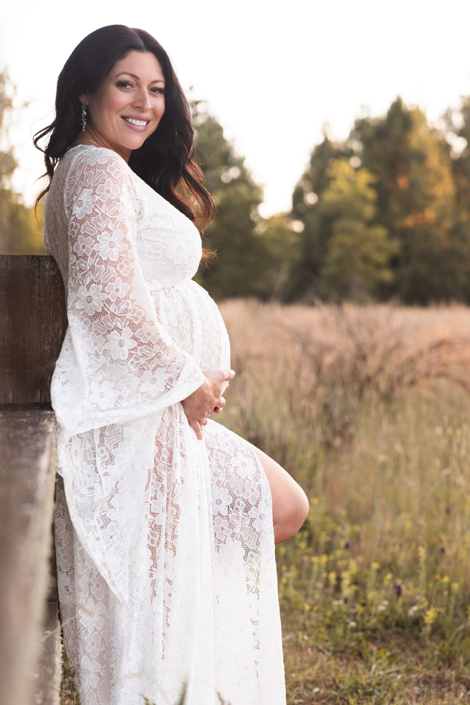 A maternity photo featuring an elegant white lace dress in a sunlit field with tall grass and trees in the background.