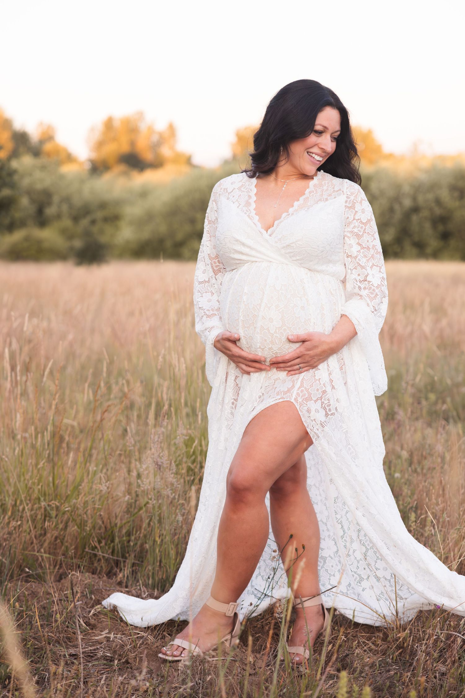 A pregnant woman in a white lace dress poses in a golden field at sunset, wearing elegant heels.
