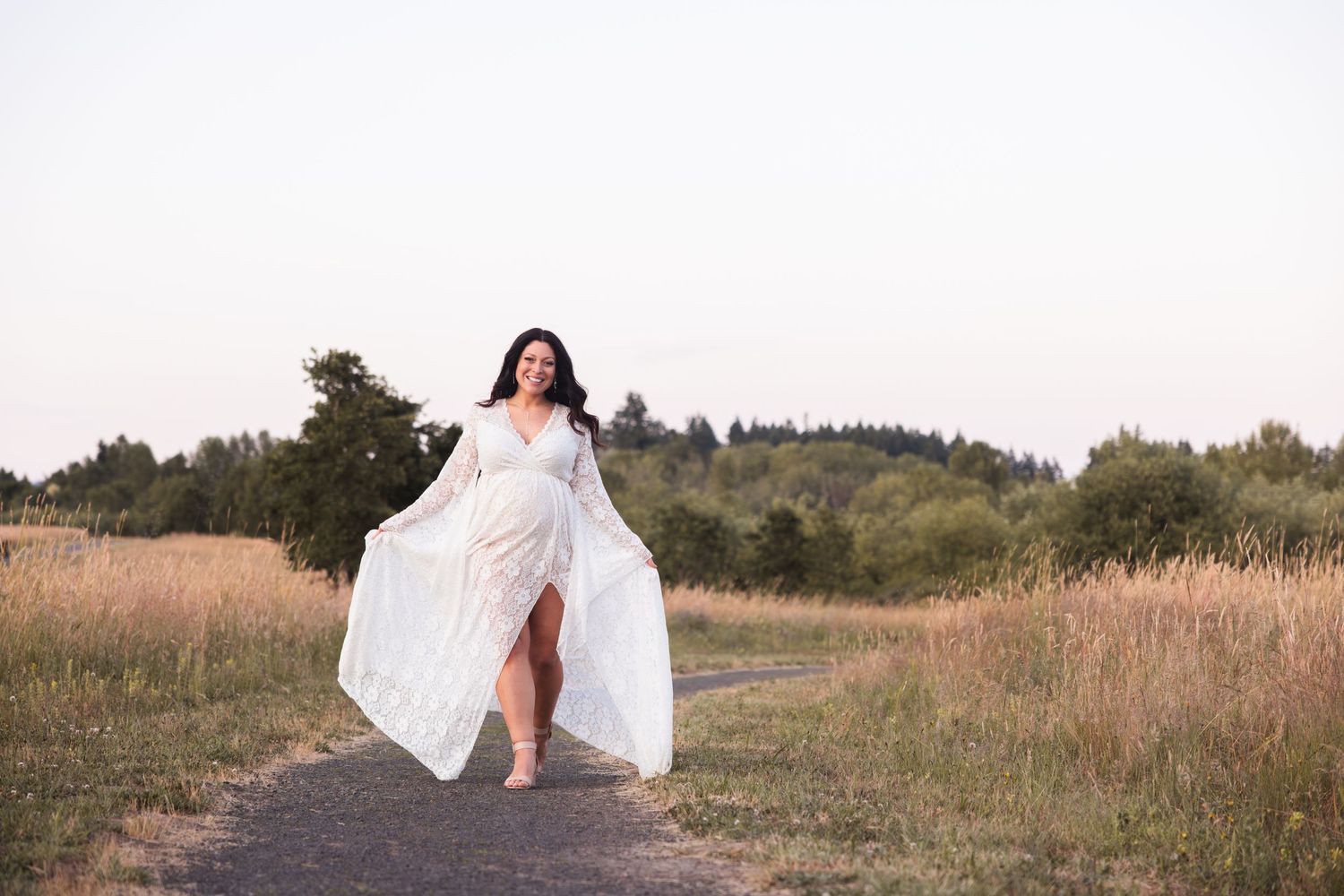 A flowing white dress flows in the breeze as a person walks along a rural dirt path at sunset.