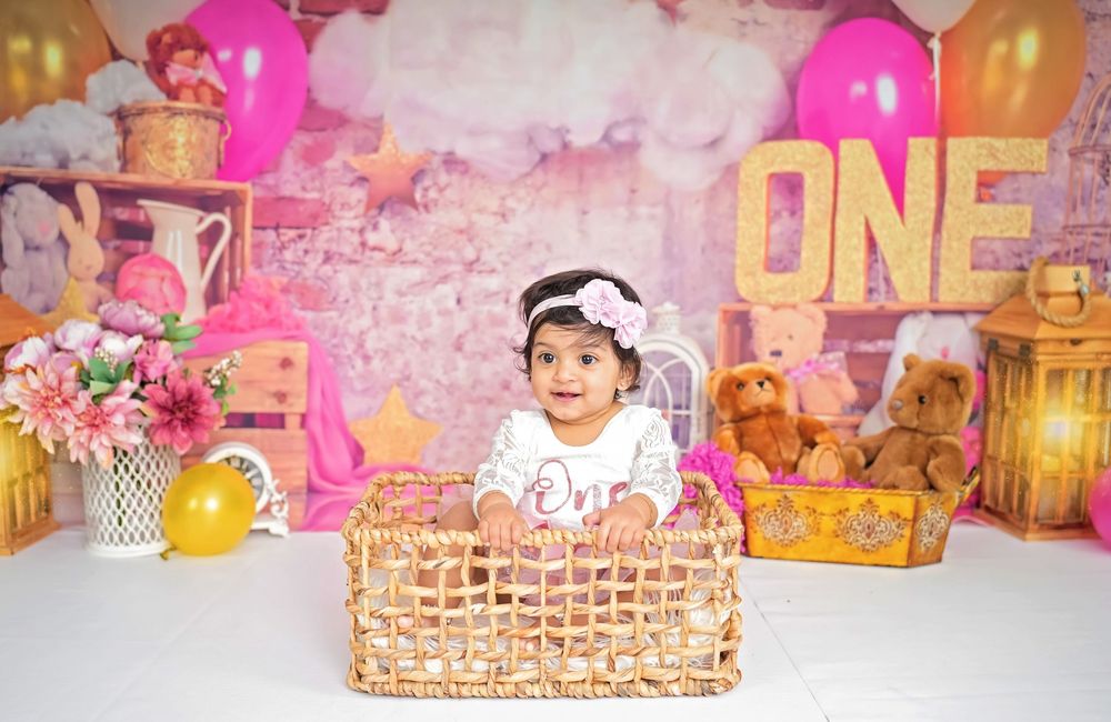 Pink and gold themed first birthday party setup with wicker baskets, balloons, and floral decorations.