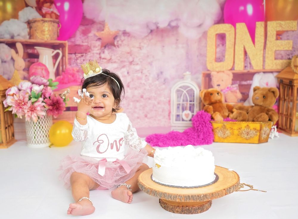 A baby in a white outfit sits by a first birthday cake with pink decorations and balloons in the background.