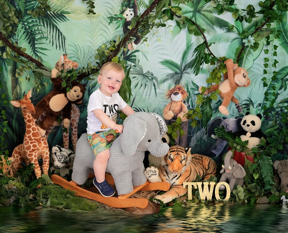 Whimsical jungle-themed photo scene with stuffed animals including elephant, giraffe, monkeys, tigers and pandas in lush greenery.