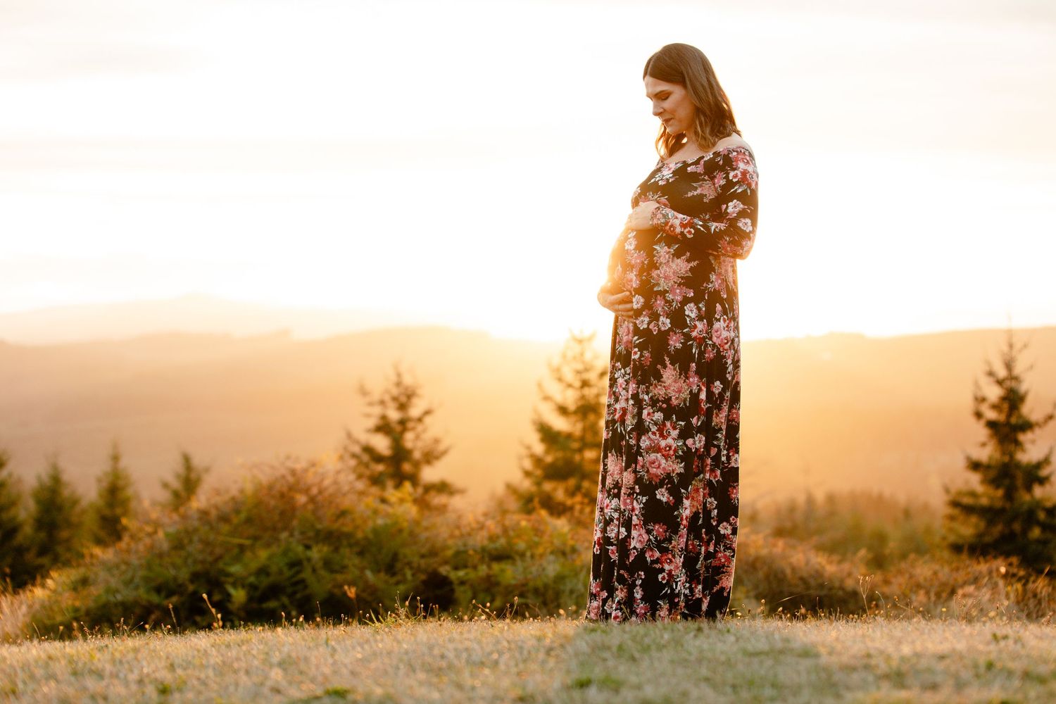 A person in a floral dress stands on a hilltop during golden hour sunset with evergreen trees in the background.