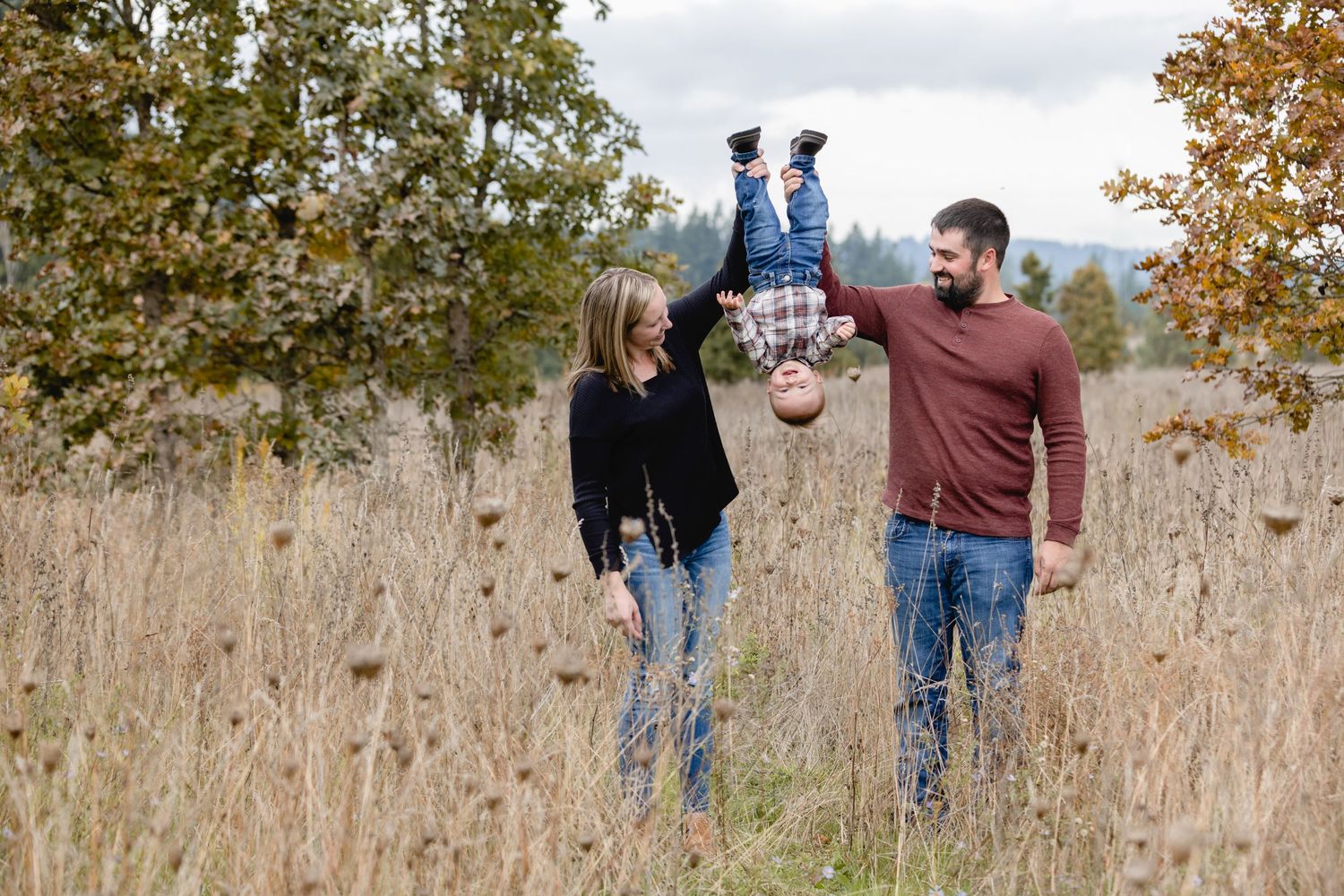 Family playfully holds small child upside down while standing in tall autumn grass with trees in background.