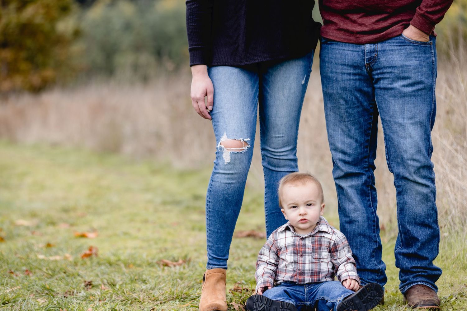 Small child in plaid shirt sits on grass between two pairs of legs in jeans on an autumn day.