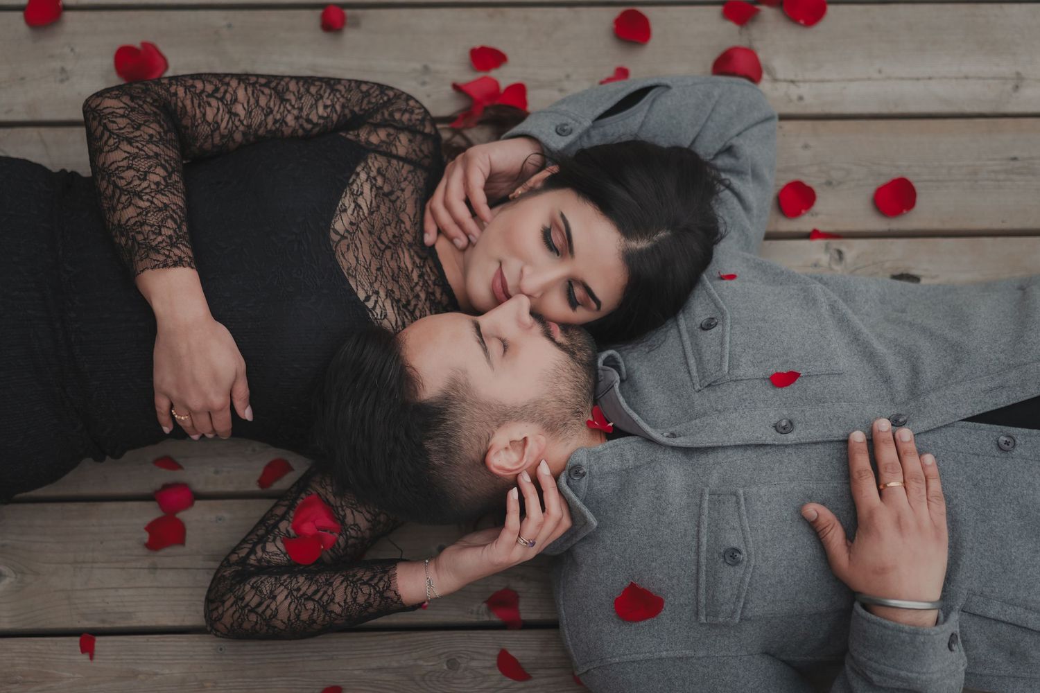 Romantic couple lying on a wooden deck surrounded by red rose petals, capturing intimate love and deep connection. Perfect for engagement