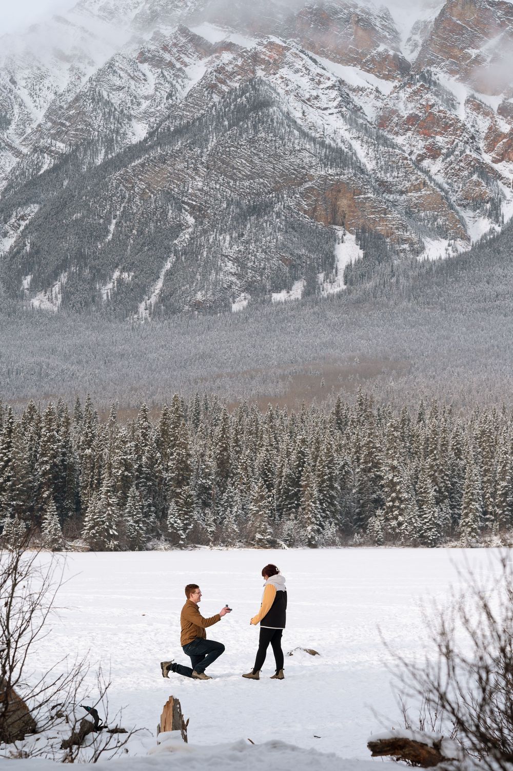 A winter proposal scene in a snowy mountain landscape with evergreen forests and majestic peaks in the background.