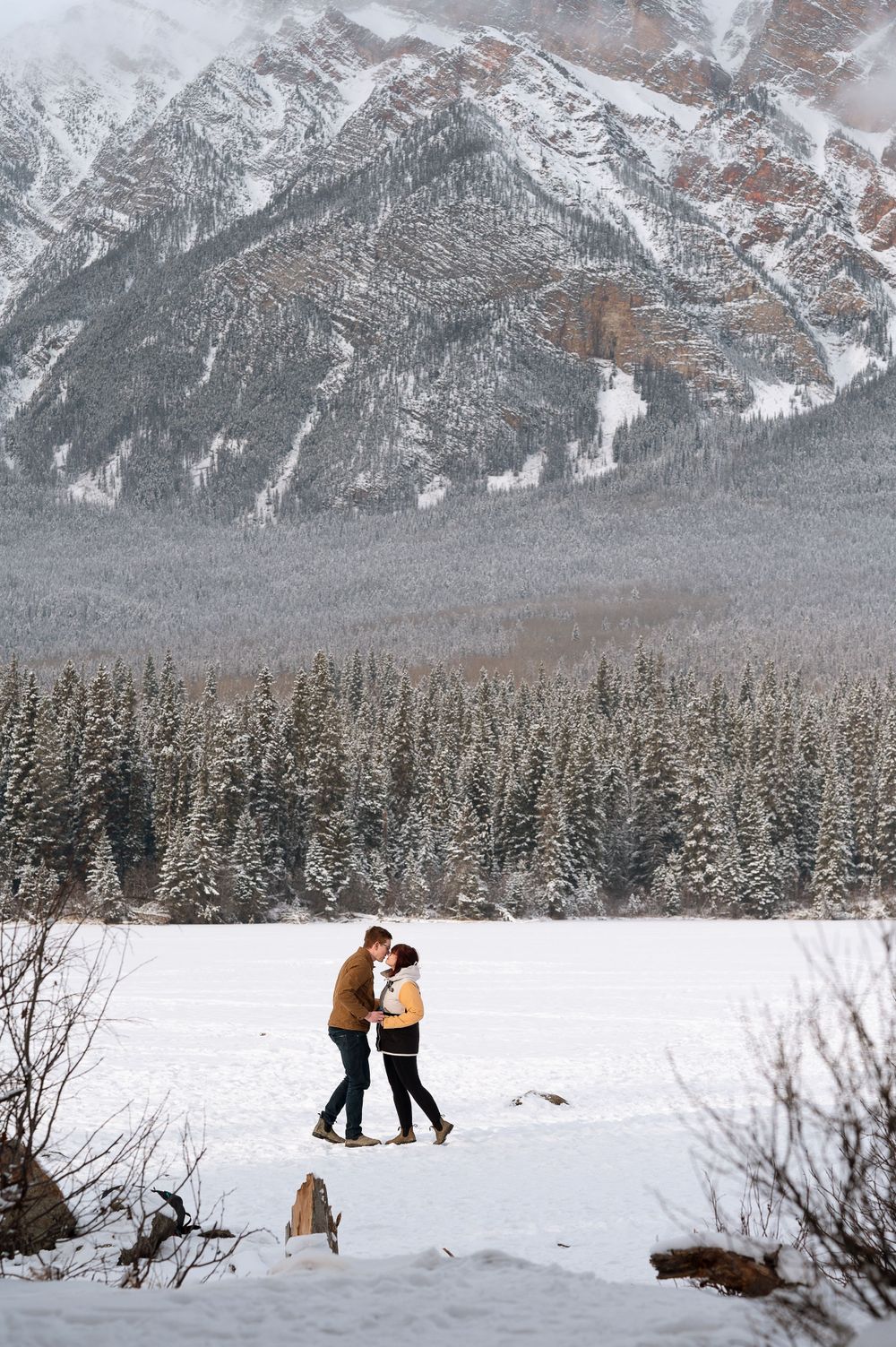 A couple embracing in winter with snow-capped mountains and pine forest in the background.