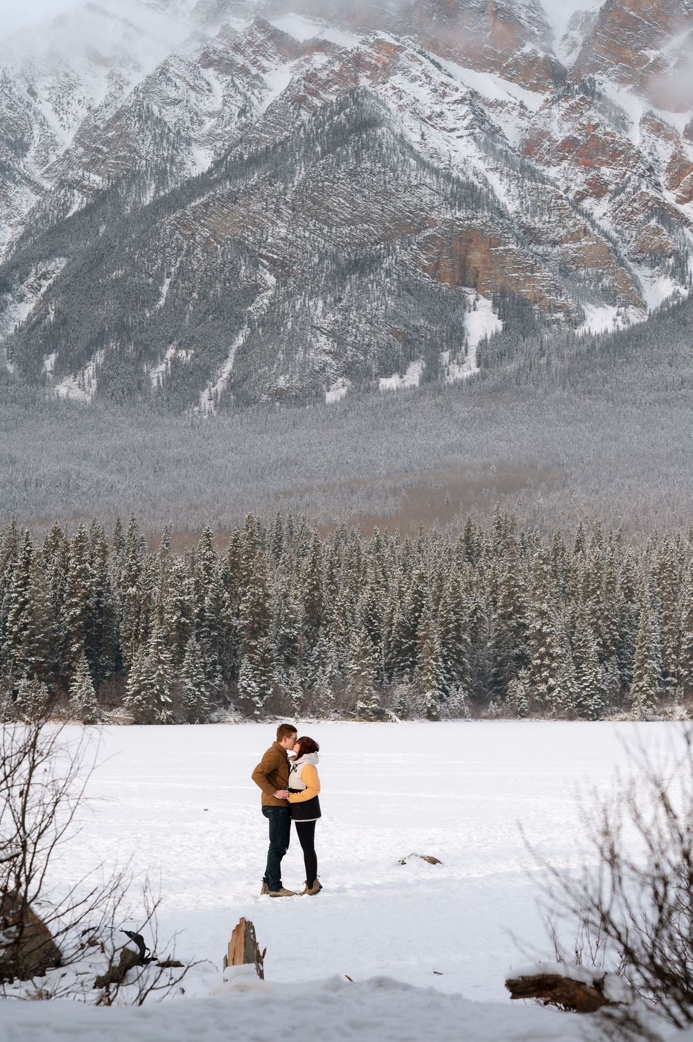 Two figures embrace in a snowy winter landscape with majestic mountains and evergreen forest in the background.