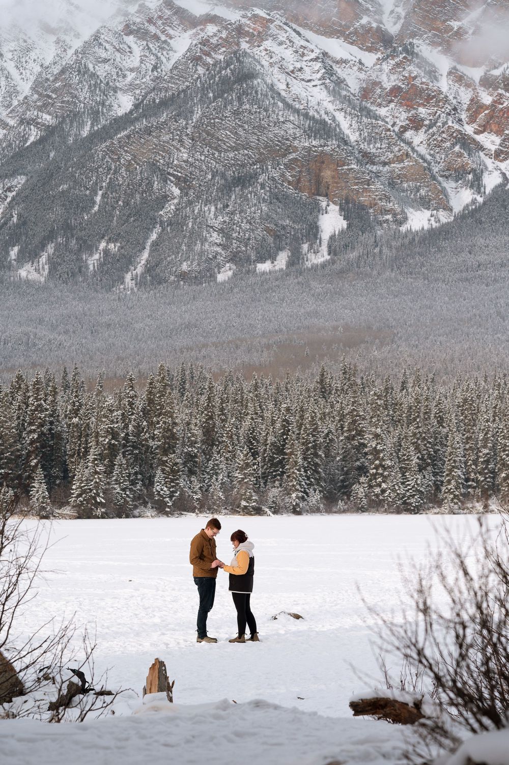 Two people standing together in a snowy field with majestic mountain peaks and forest scenery.