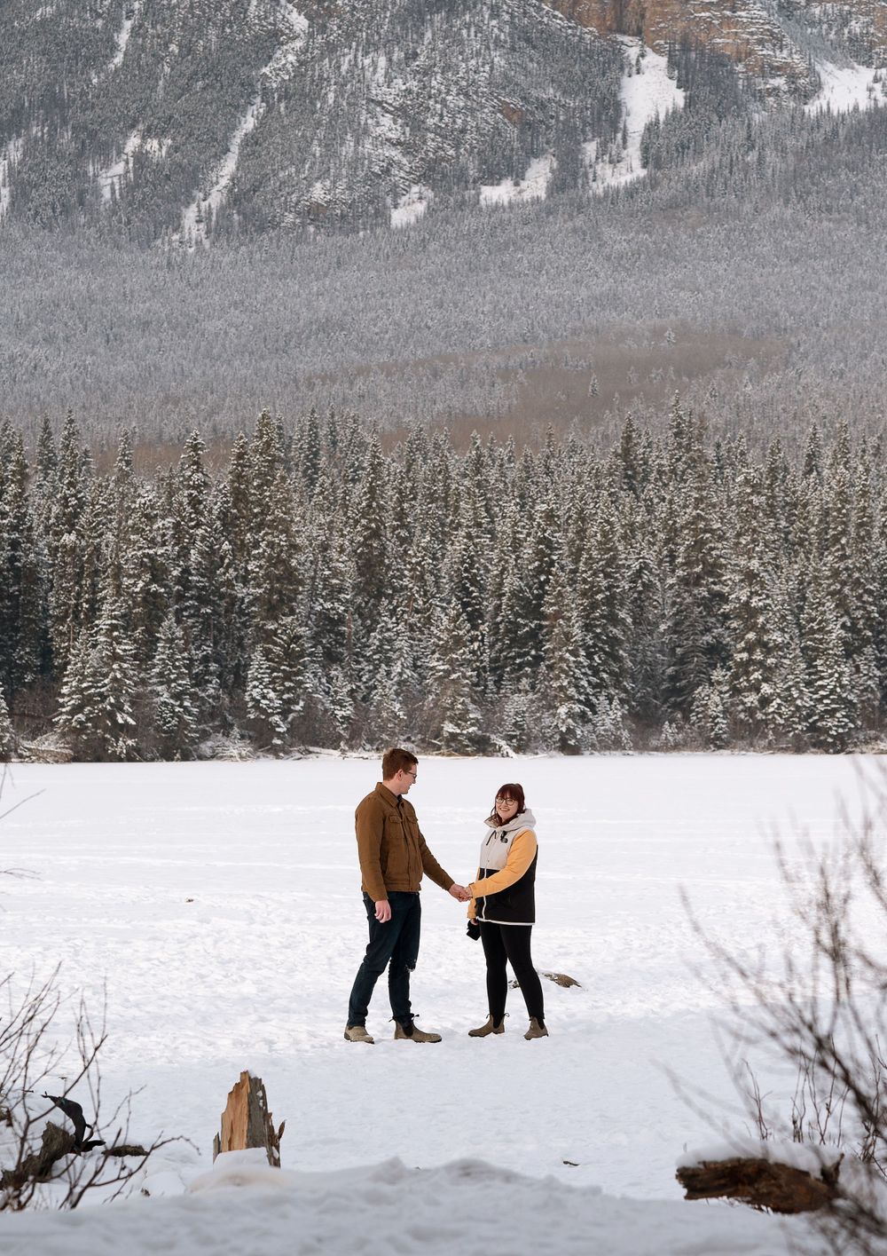 A couple stands together in a snowy winter landscape with snow-covered evergreen forest and mountains in the background.