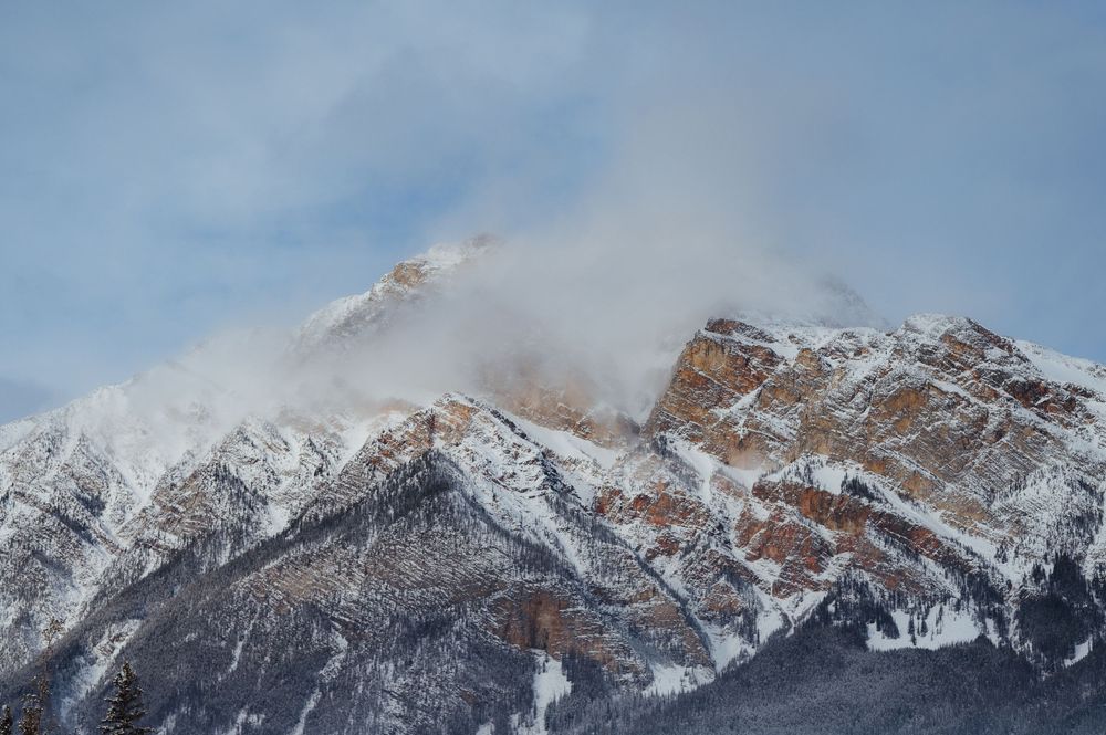 Snow-capped mountain peak with clouds swirling around its rocky cliffs against a blue sky in the Canadian Rockies.