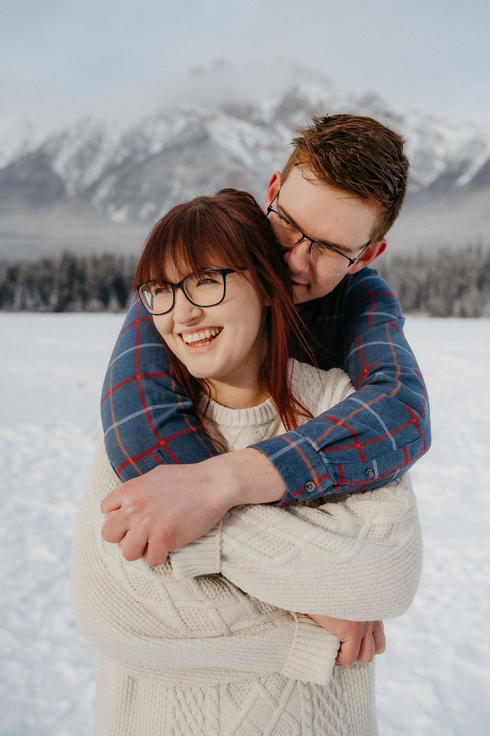 A couple embraces and laughs together on a snowy winter day with mountains behind them.