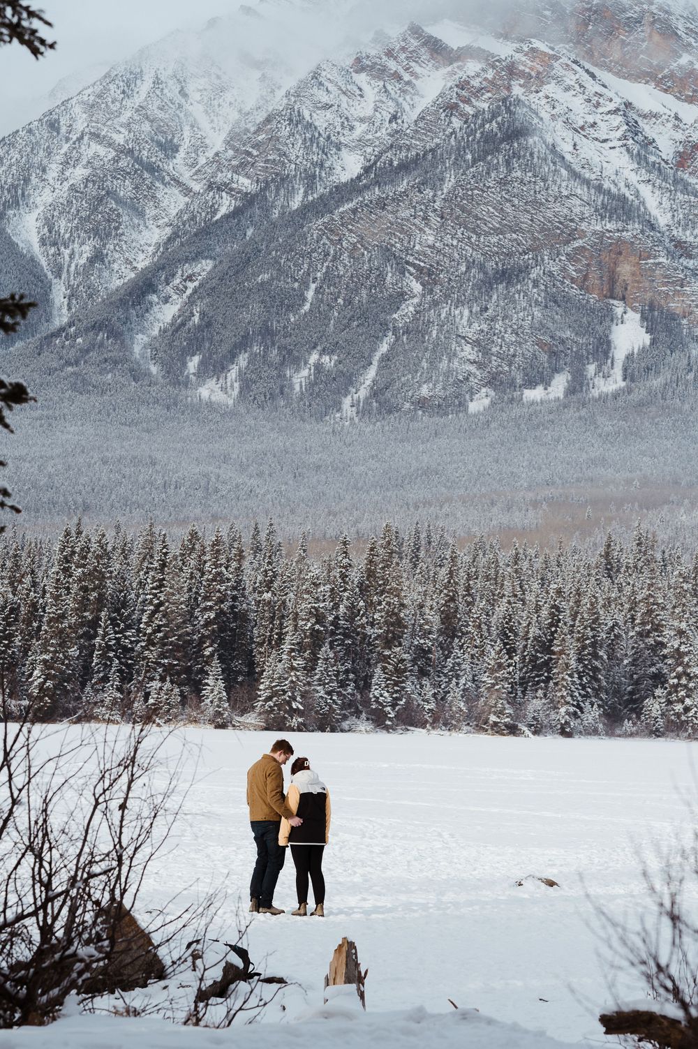 Two figures stand together in a snowy landscape with dramatic mountain peaks and evergreen forest backdrop.