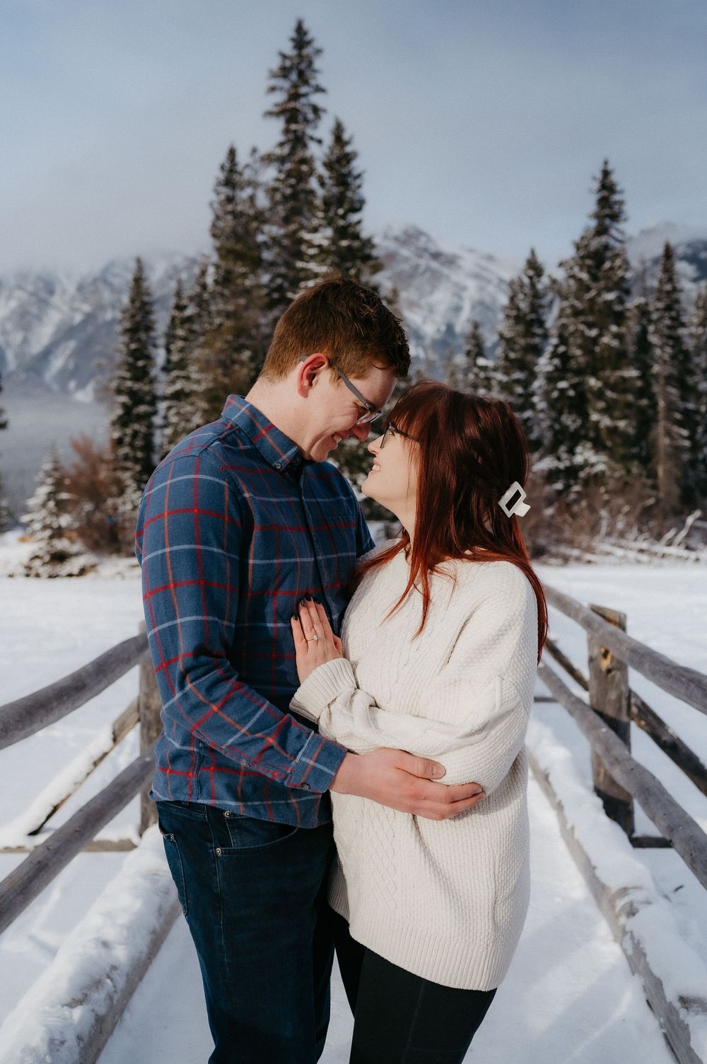 A romantic winter embrace on a snow-covered wooden bridge with mountain scenery in the background.