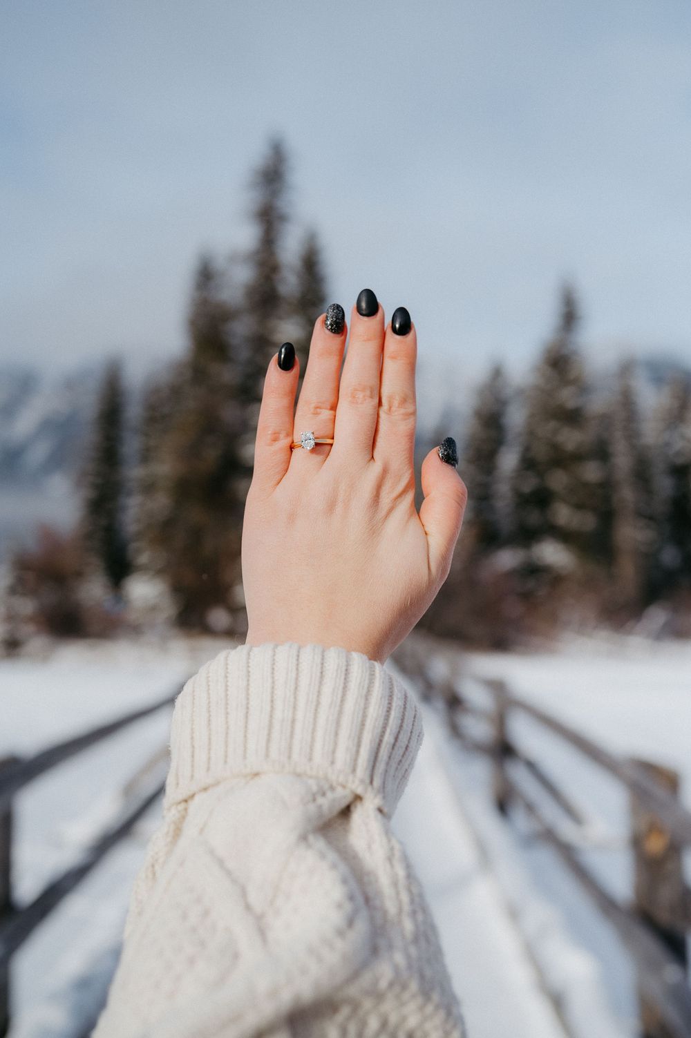 A hand wearing a white knit sweater shows off dark nail polish and an engagement ring against a snowy winter landscape.